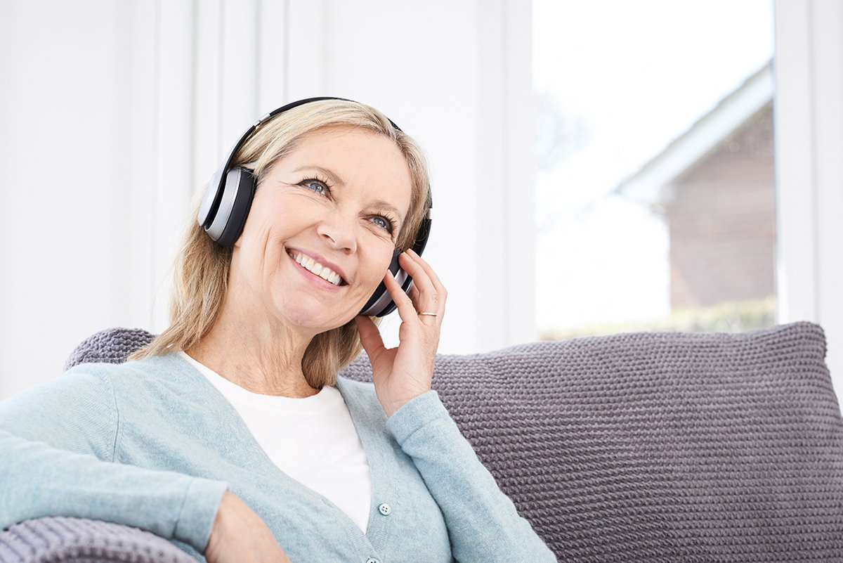 Ältere Frau sitzt auf dem Sofa und hört Musik über Kopfhörer